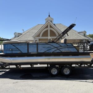 Blue and silver 2026 Sylvan Mirage X3 CLZ DH tritoon on a tandem-axle trailer, with the outboard and raised bimini visible